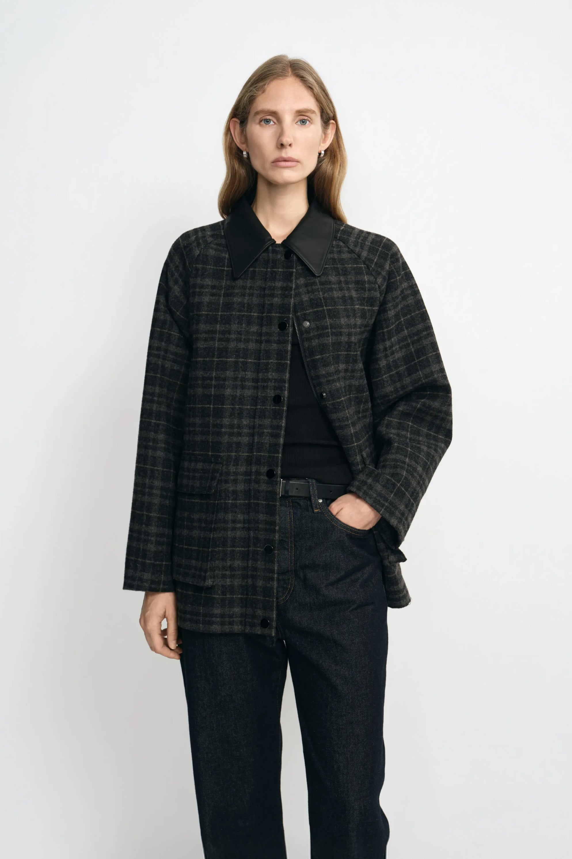 A model wearing a grey plaid wool-blend jacket with a contrasting black sheepskin collar, layered over a black top and dark jeans.