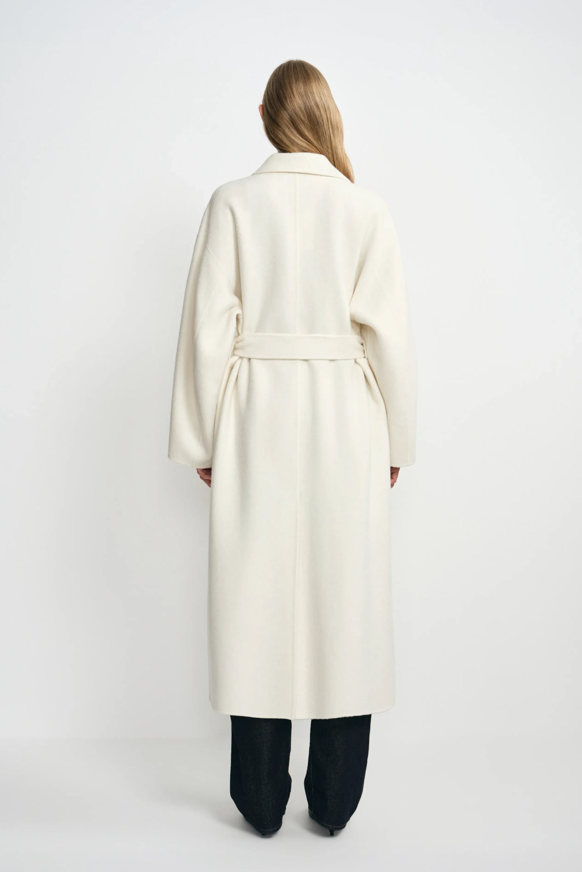 Back view of a model wearing a winter-white wool coat, with the belt gathering the waist above a long hem and dark jeans.
