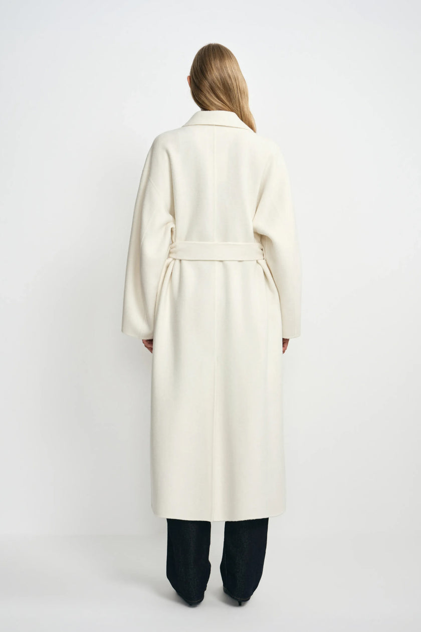 Back view of a model wearing a winter-white wool coat, with the belt gathering the waist above a long hem and dark jeans.