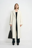 A model facing forward in an open winter-white wool coat with large patch pockets, paired with a black top, dark jeans, black shoes and a large black bag.