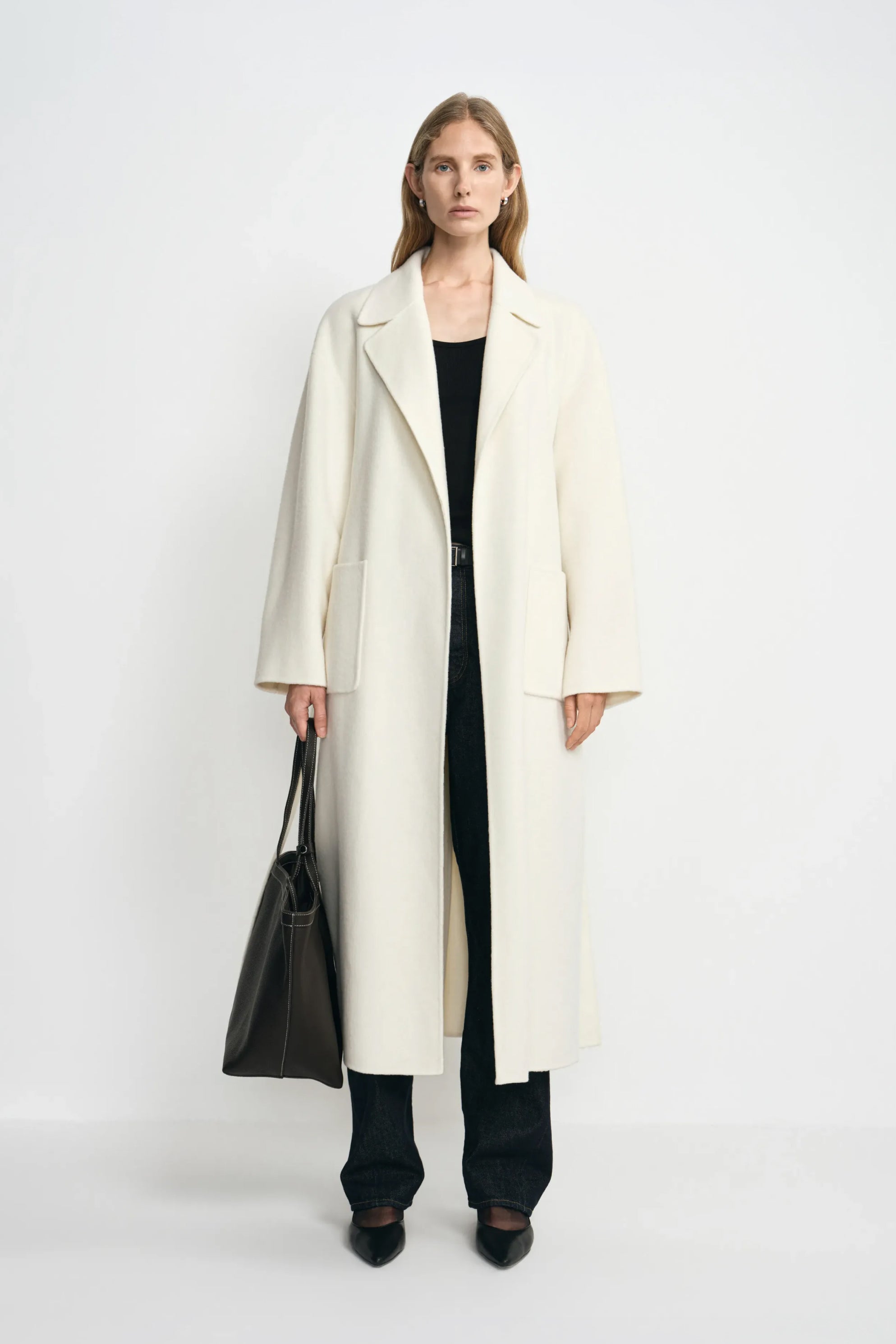 A model facing forward in an open winter-white wool coat with large patch pockets, paired with a black top, dark jeans, black shoes and a large black bag.