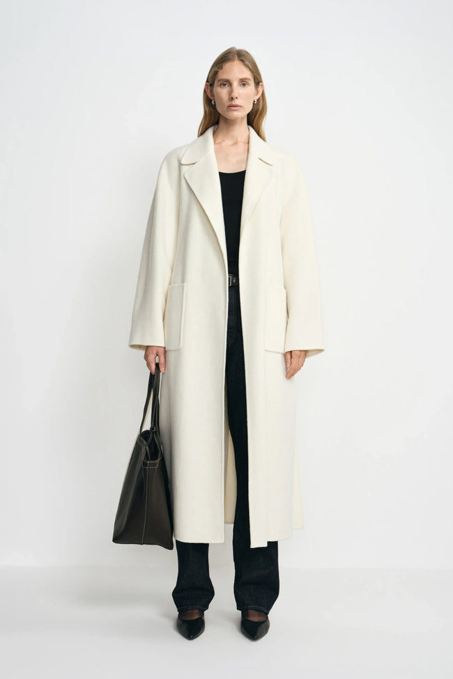 A model facing forward in an open winter-white wool coat with large patch pockets, paired with a black top, dark jeans, black shoes and a large black bag.