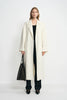 A model facing forward in an open winter-white wool coat with large patch pockets, paired with a black top, dark jeans, black shoes and a large black bag.