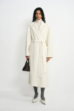 A model wearing a belted winter-white wool coat with broad lapels, styled with a cream turtleneck, grey socks, pointed white shoes and a small dark bag.
