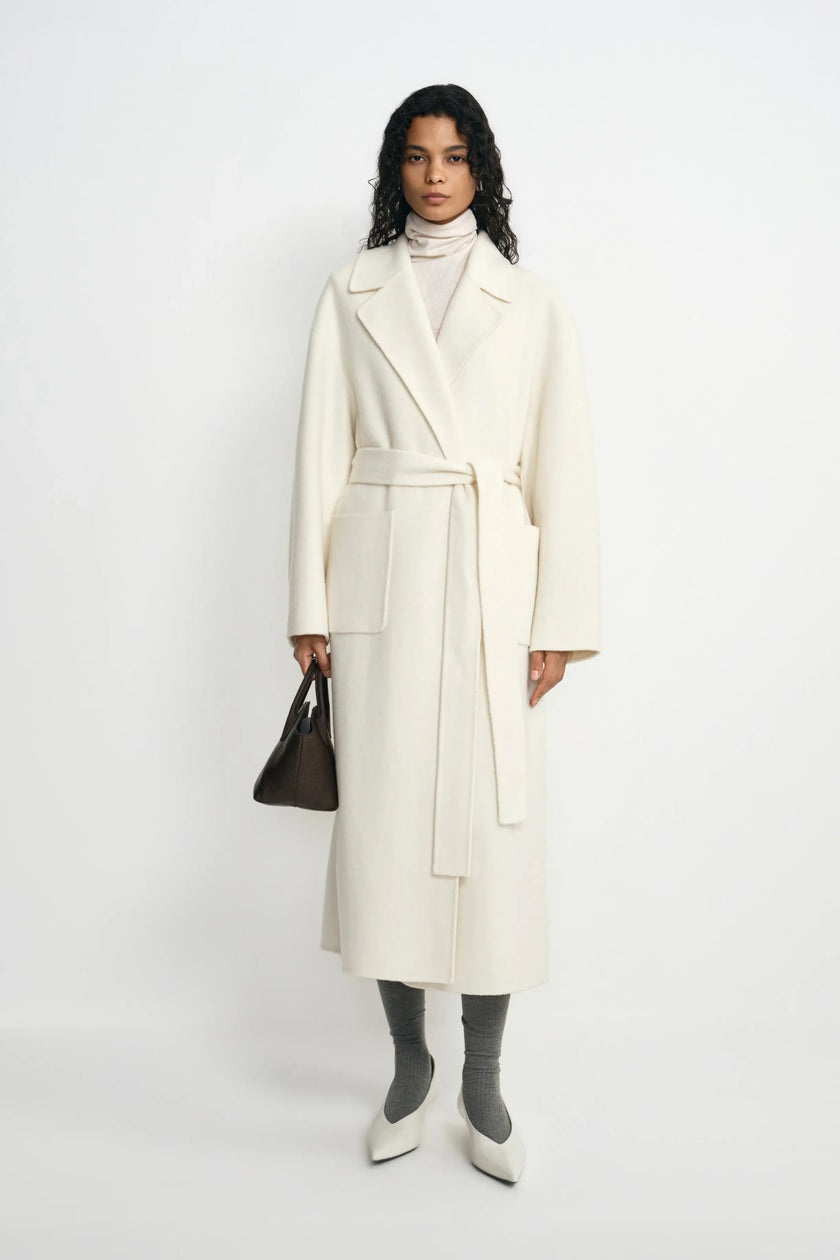 A model wearing a belted winter-white wool coat with broad lapels, styled with a cream turtleneck, grey socks, pointed white shoes and a small dark bag.