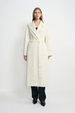 A model wearing a winter-white 100% wool wrap coat with notched lapels and a tied waist, paired with a black top, dark jeans and pointed black shoes.