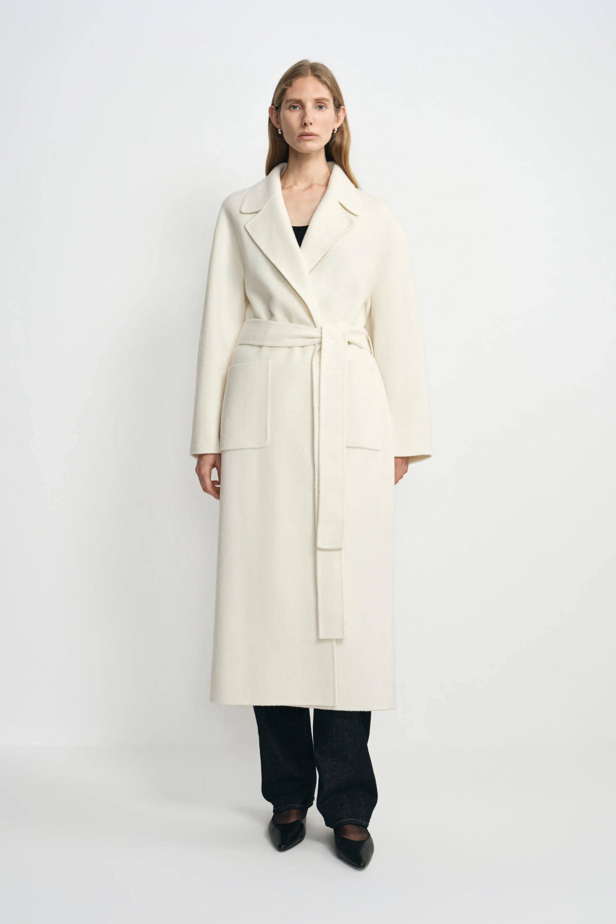 A model wearing a winter-white 100% wool wrap coat with notched lapels and a tied waist, paired with a black top, dark jeans and pointed black shoes.