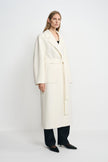 Three-quarter view of a model in a winter-white wool coat, showing the wrap front, tied belt and large patch pocket above dark jeans and black shoes.