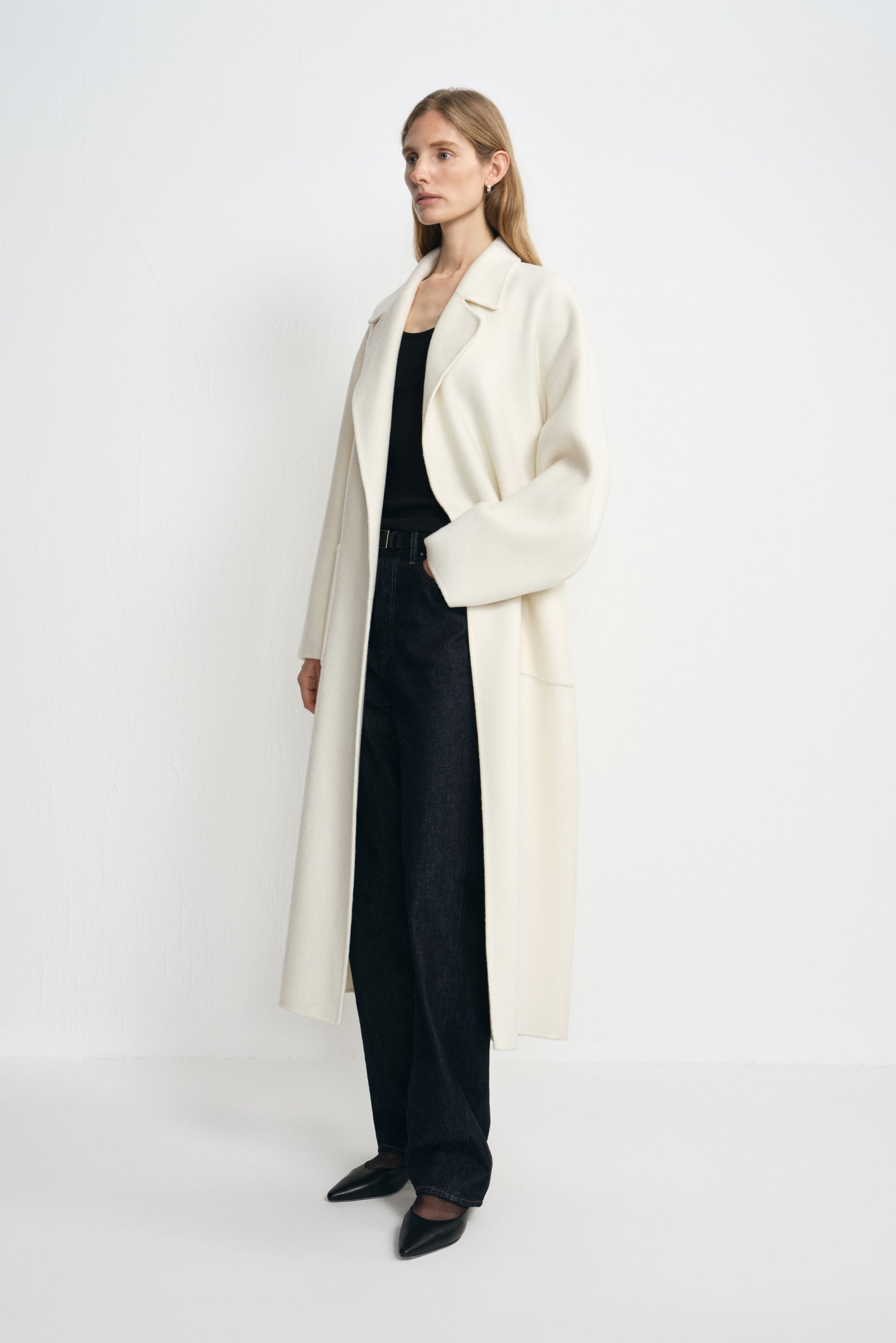 Side view of a model in an open winter-white wool coat, with one hand in a patch pocket, wearing a black tank, dark jeans and pointed shoes.