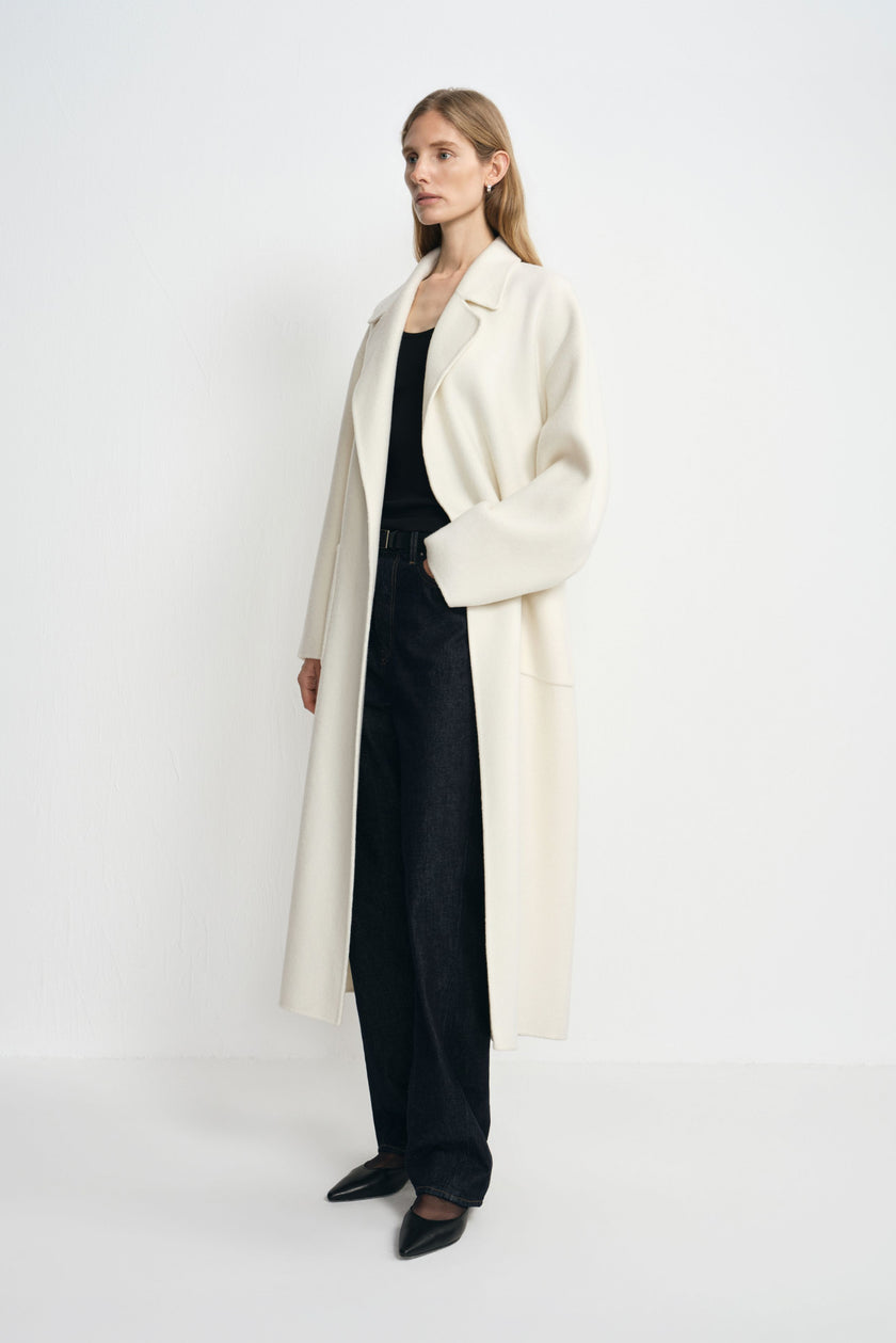 Side view of a model in an open winter-white wool coat, with one hand in a patch pocket, wearing a black tank, dark jeans and pointed shoes.