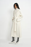 Three-quarter view of a model in a belted winter-white wool coat with large patch pockets, paired with a cream turtleneck, grey socks and white pointed shoes.