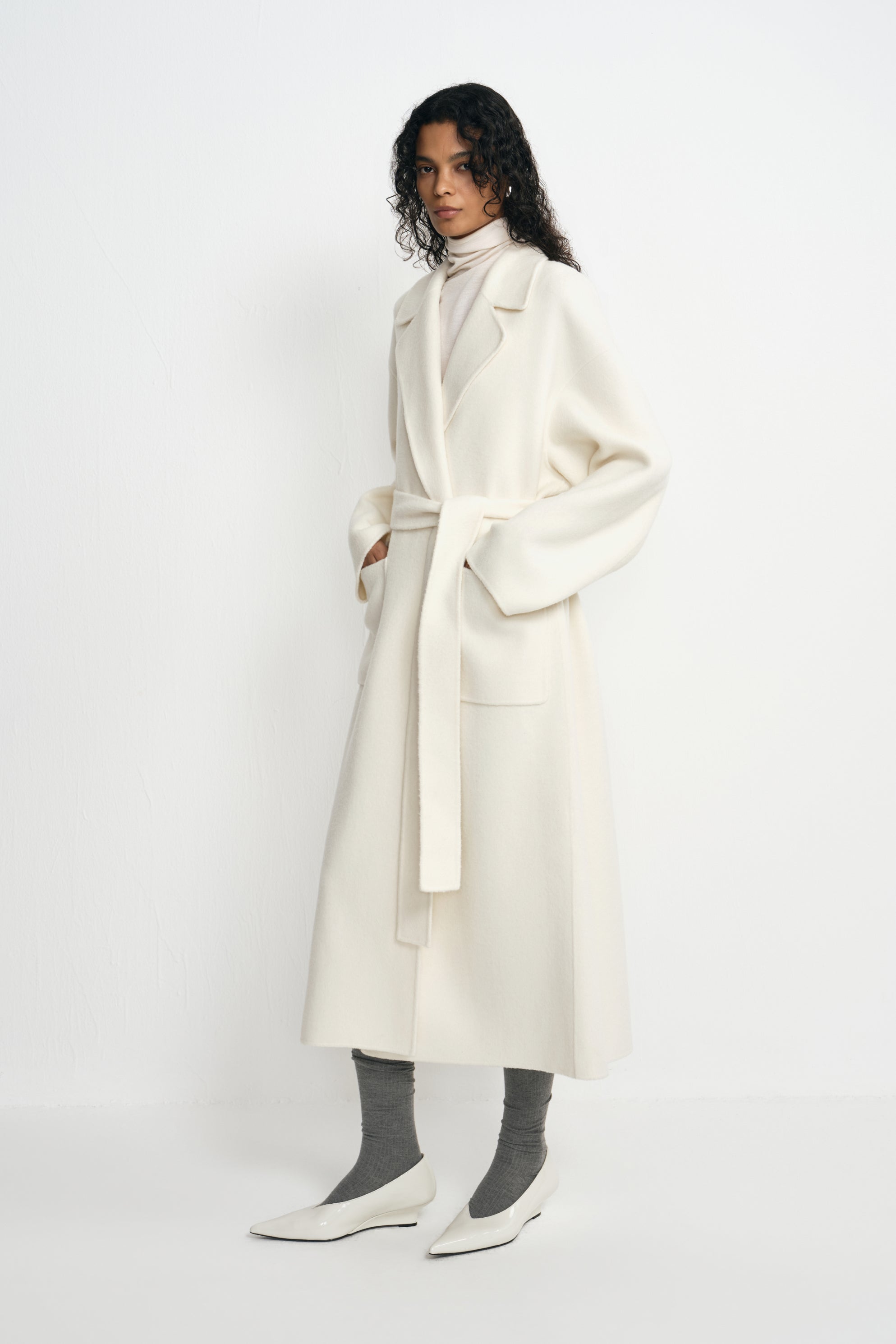 Three-quarter view of a model in a belted winter-white wool coat with large patch pockets, paired with a cream turtleneck, grey socks and white pointed shoes.