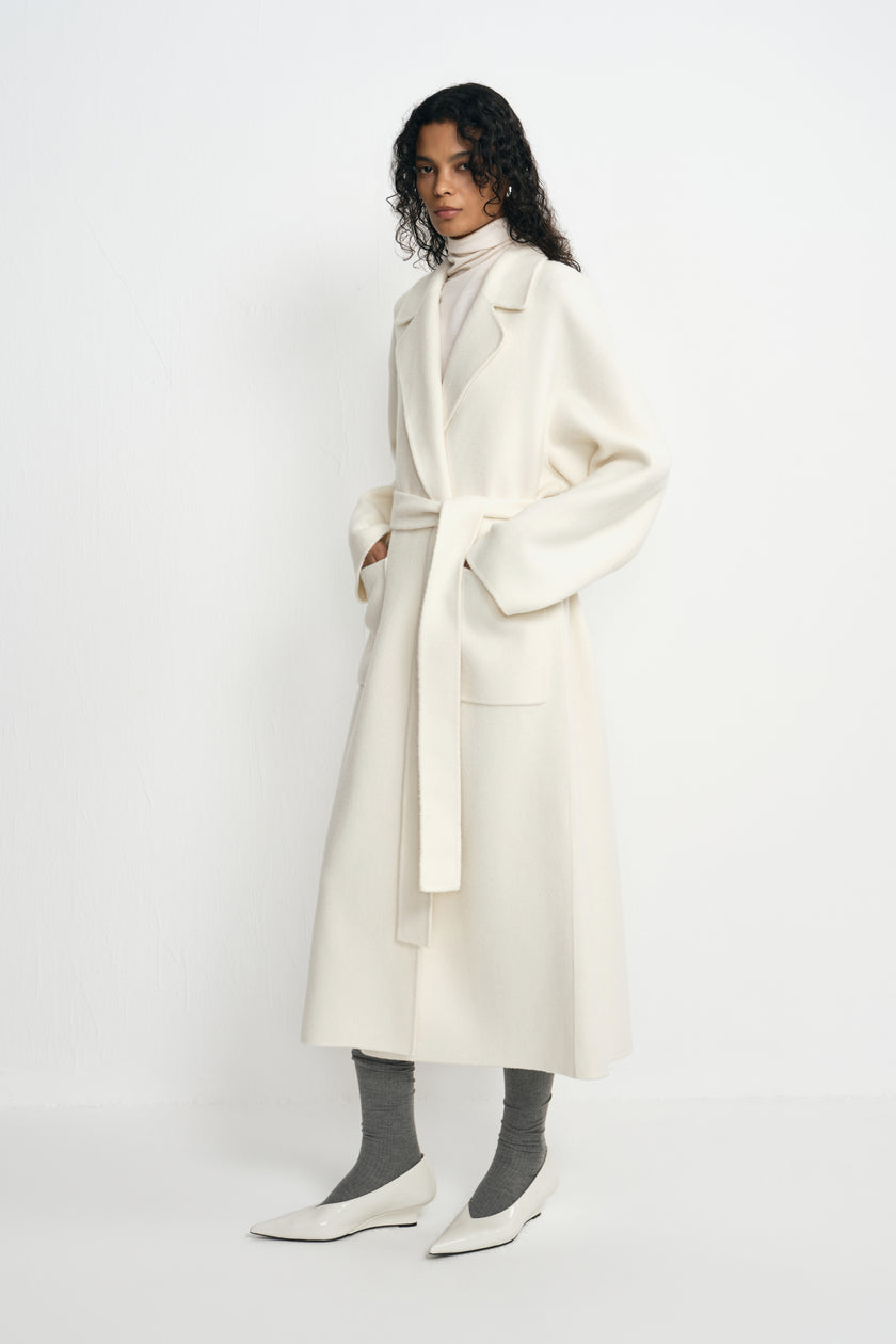 Three-quarter view of a model in a belted winter-white wool coat with large patch pockets, paired with a cream turtleneck, grey socks and white pointed shoes.