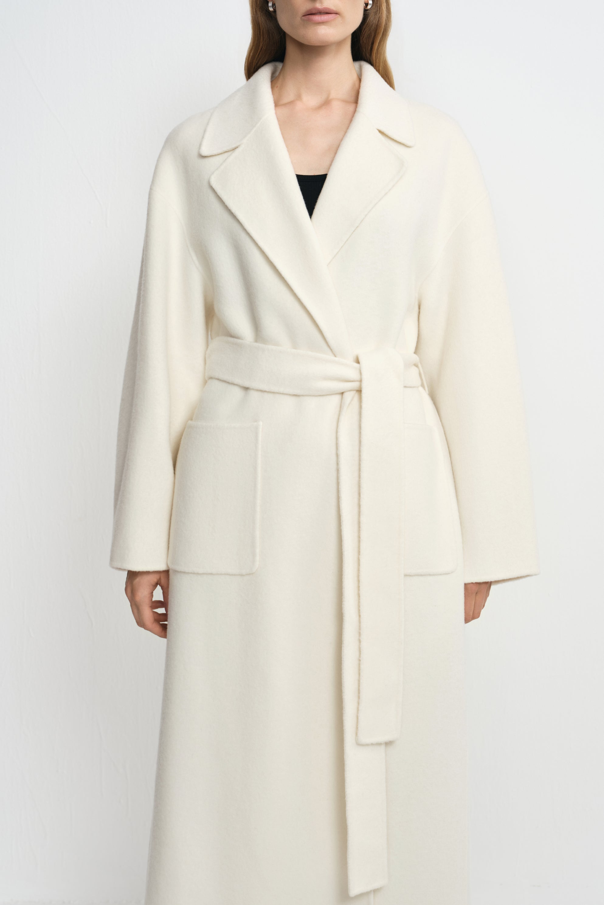 Close-up of a winter-white 100% wool wrap coat on a model, showing broad notched lapels, a tied waist belt and large rectangular patch pockets.