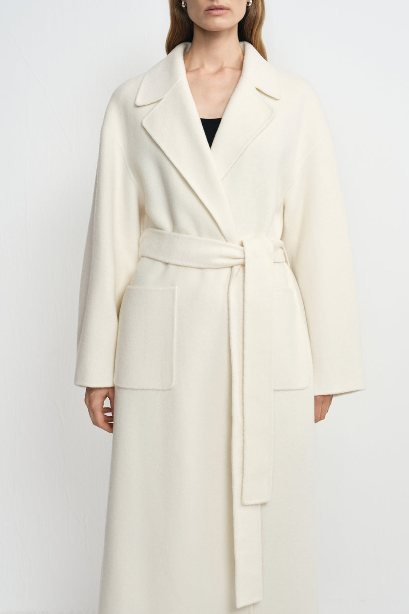 Close-up of a winter-white 100% wool wrap coat on a model, showing broad notched lapels, a tied waist belt and large rectangular patch pockets.