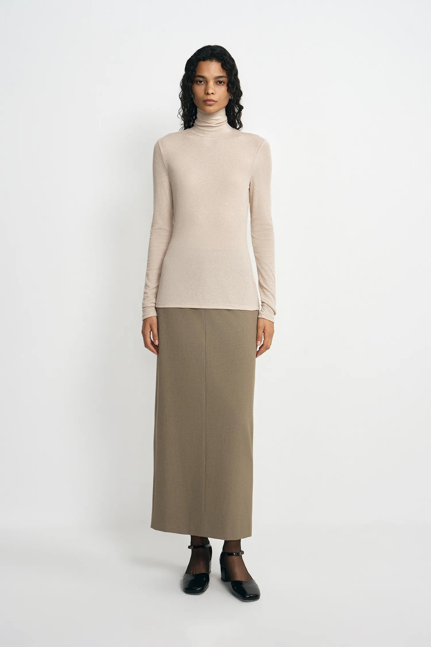 A model wearing an oatmeal pencil midi skirt with a slim tailored fit, pairing a beige turtleneck top and black Mary Jane heels.
