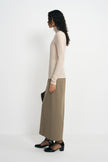 A woman stands in profile wearing a fitted cream turtleneck sweater and a long taupe skirt, paired with black shoes, against a plain light gray studio background.