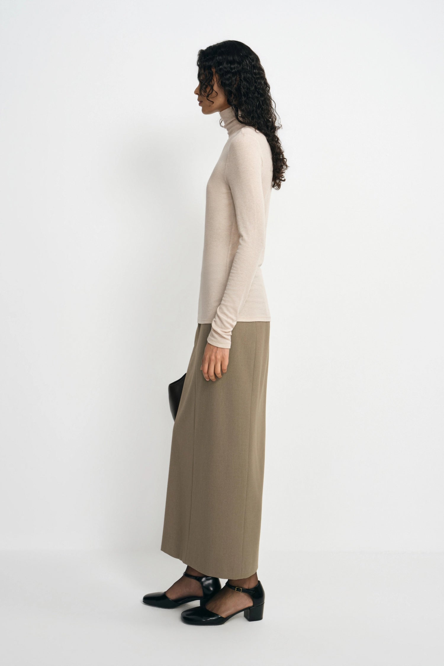 A woman stands in profile wearing a fitted cream turtleneck sweater and a long taupe skirt, paired with black shoes, against a plain light gray studio background.