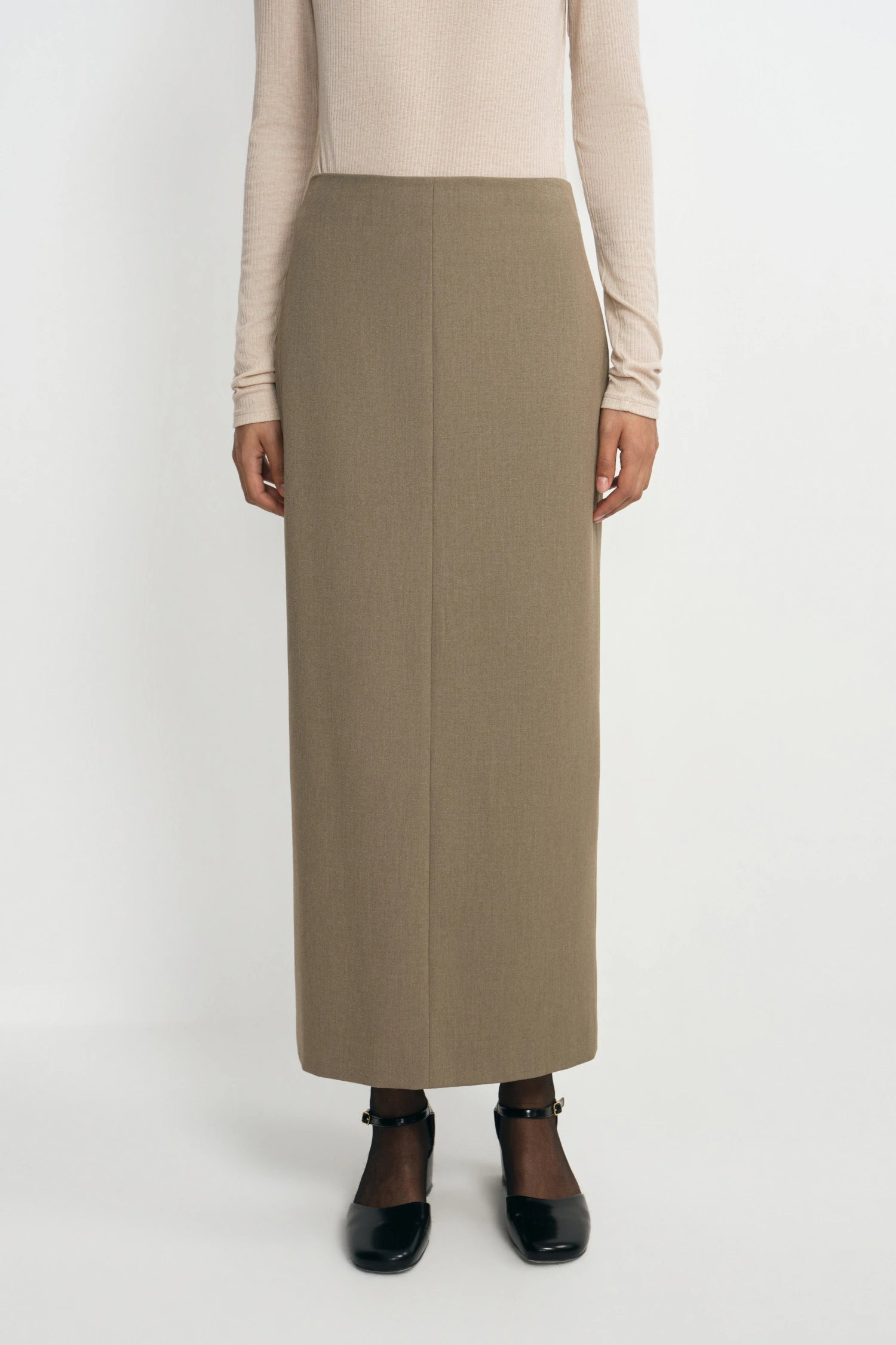A model is shown from the chest down wearing a long, straight, ankle-length taupe skirt, a beige long-sleeve top, and black Mary Jane shoes against a plain white studio background.