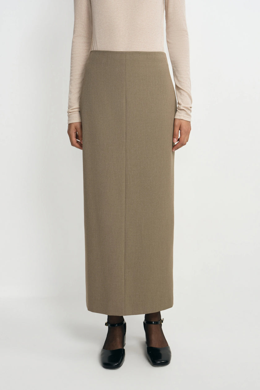 A model is shown from the chest down wearing a long, straight, ankle-length taupe skirt, a beige long-sleeve top, and black Mary Jane shoes against a plain white studio background.