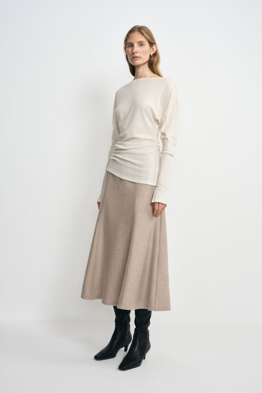 Three-quarter view of a model in an oatmeal wool midi skirt, with the flared hem below an untucked cream top and above black boots.