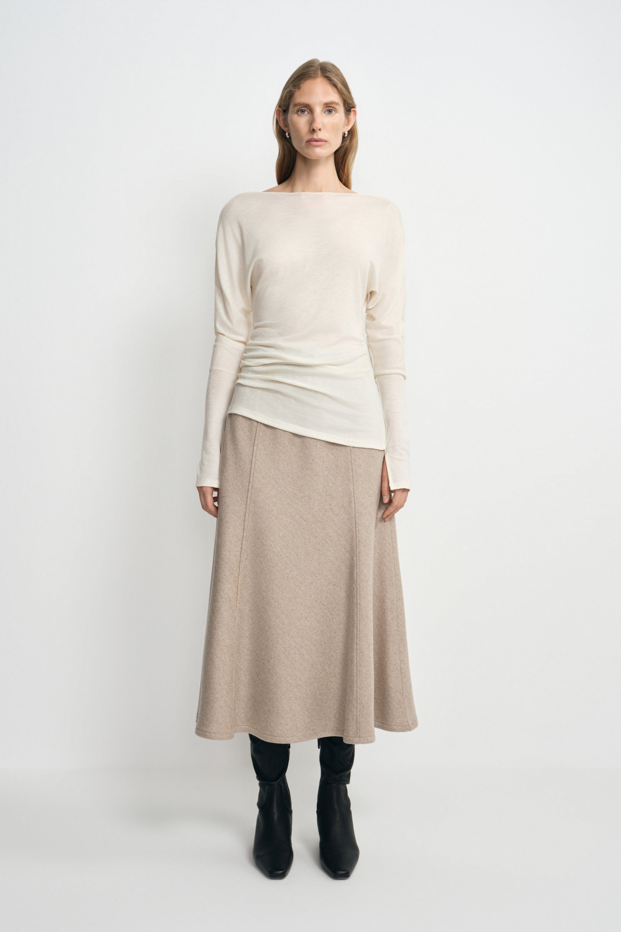 A model facing forward in an oatmeal wool midi skirt with an A-line silhouette, styled with a cream long-sleeve top and black boots.