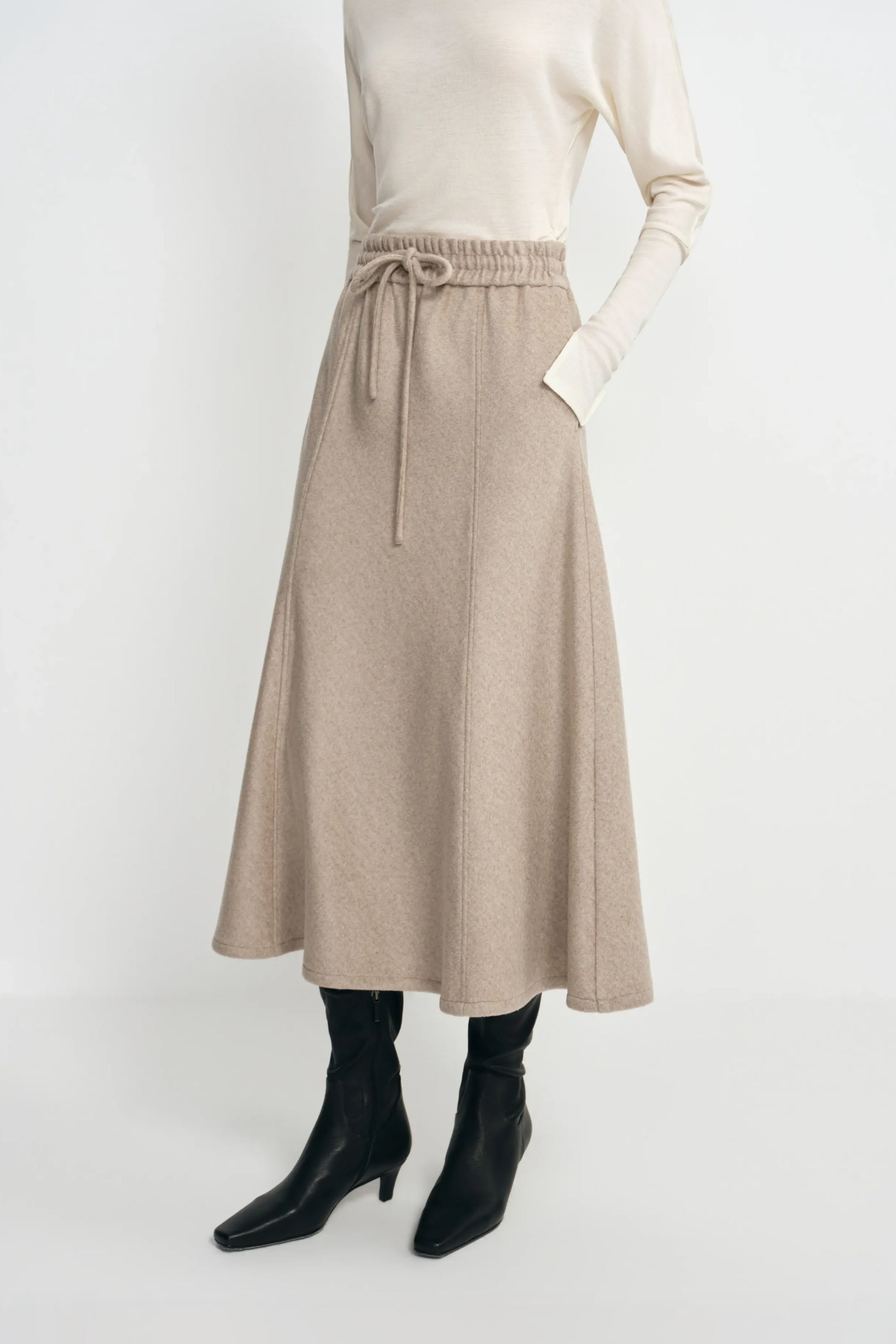 Close-up of an oatmeal wool midi skirt on a model, showing the gathered drawstring waist and flared hem, paired with a cream top and black boots.