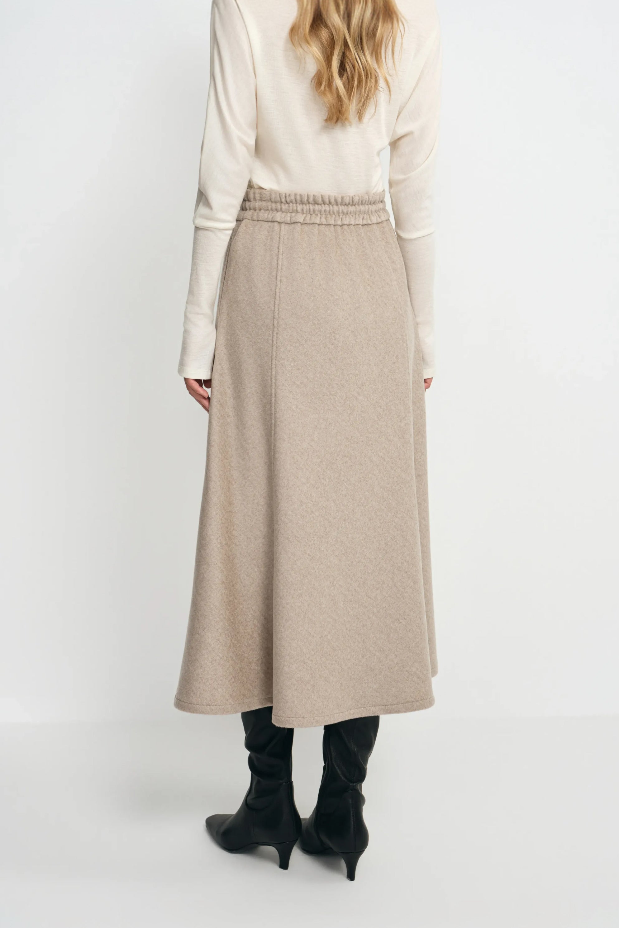 Rear three-quarter view of an oatmeal wool midi skirt on a model, showing the gathered waistband above a flared hem and black heeled boots.