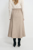 Back view of a model wearing an oatmeal wool midi skirt with a gathered waist and widening hem, paired with a tucked cream top and black boots.