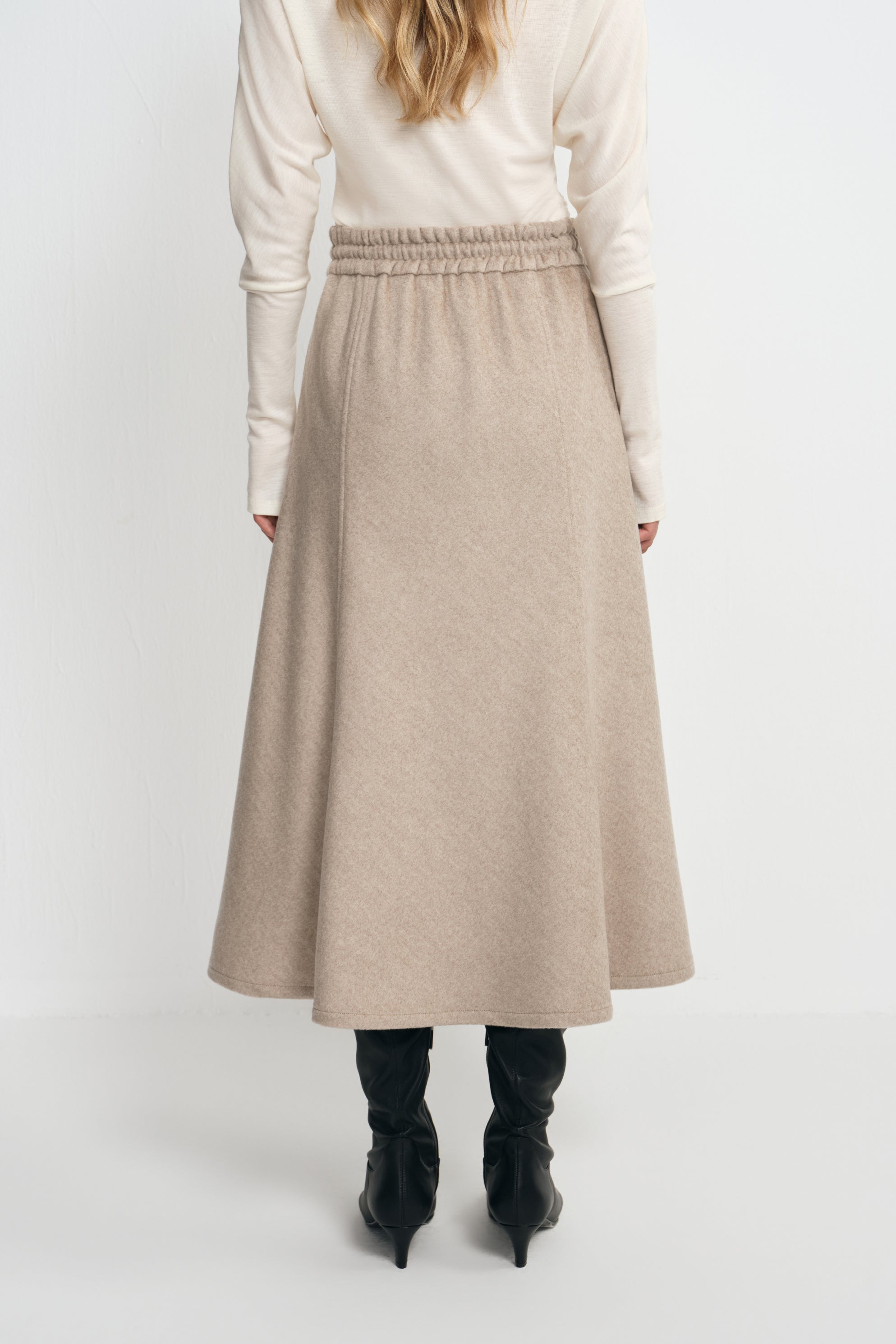 Back view of a model wearing an oatmeal wool midi skirt with a gathered waist and widening hem, paired with a tucked cream top and black boots.