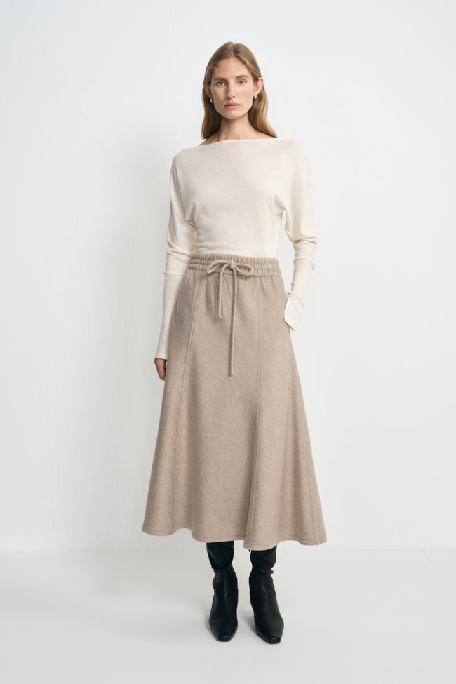 A model wearing an oatmeal 100% wool midi skirt with long waist ties and a flared hem, paired with a tucked cream top and black heeled boots.