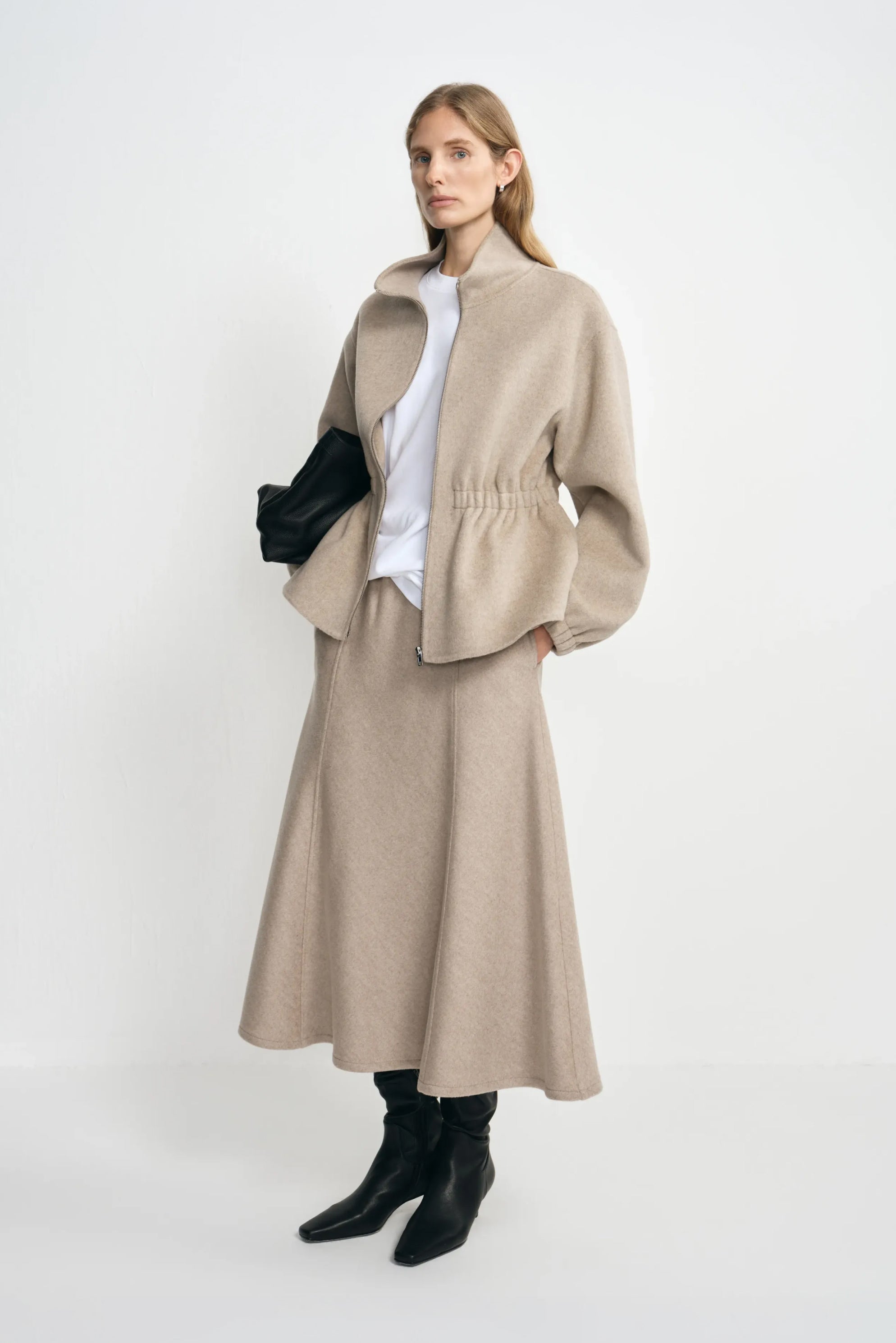 Angled view of a model in an oatmeal wool midi skirt, styled with a beige gathered-waist jacket, white T-shirt, black clutch and black boots.