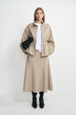 A model wearing an oatmeal 100% wool midi skirt with a flared hem, paired with a beige jacket, white T-shirt and black heeled boots.
