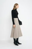 A model wearing a taupe bias-cut wool midi skirt with soft swing volume, pairing a black turtleneck top and black pointed-toe boots.
