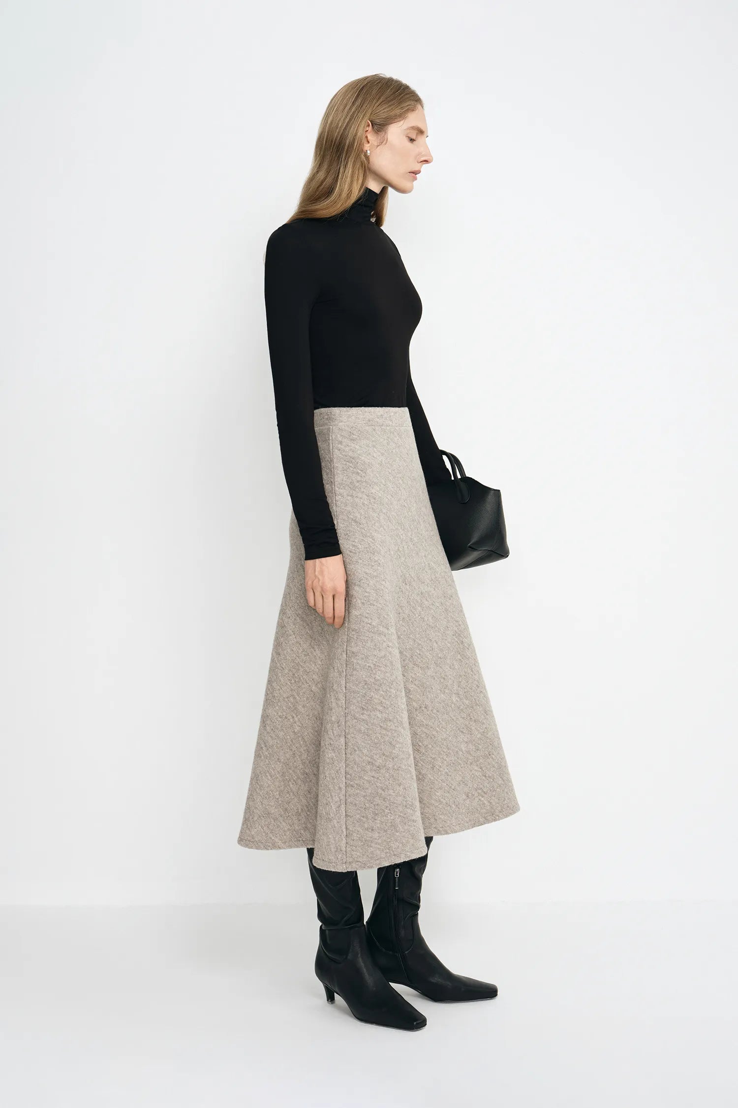 A model wearing a taupe bias-cut wool midi skirt with soft swing volume, pairing a black turtleneck top and black pointed-toe boots.