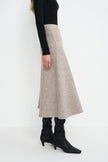 A model is shown from the side wearing a long, flared A-line midi skirt in a light oatmeal beige color, styled with a black long-sleeve top and black pointed-toe ankle boots.