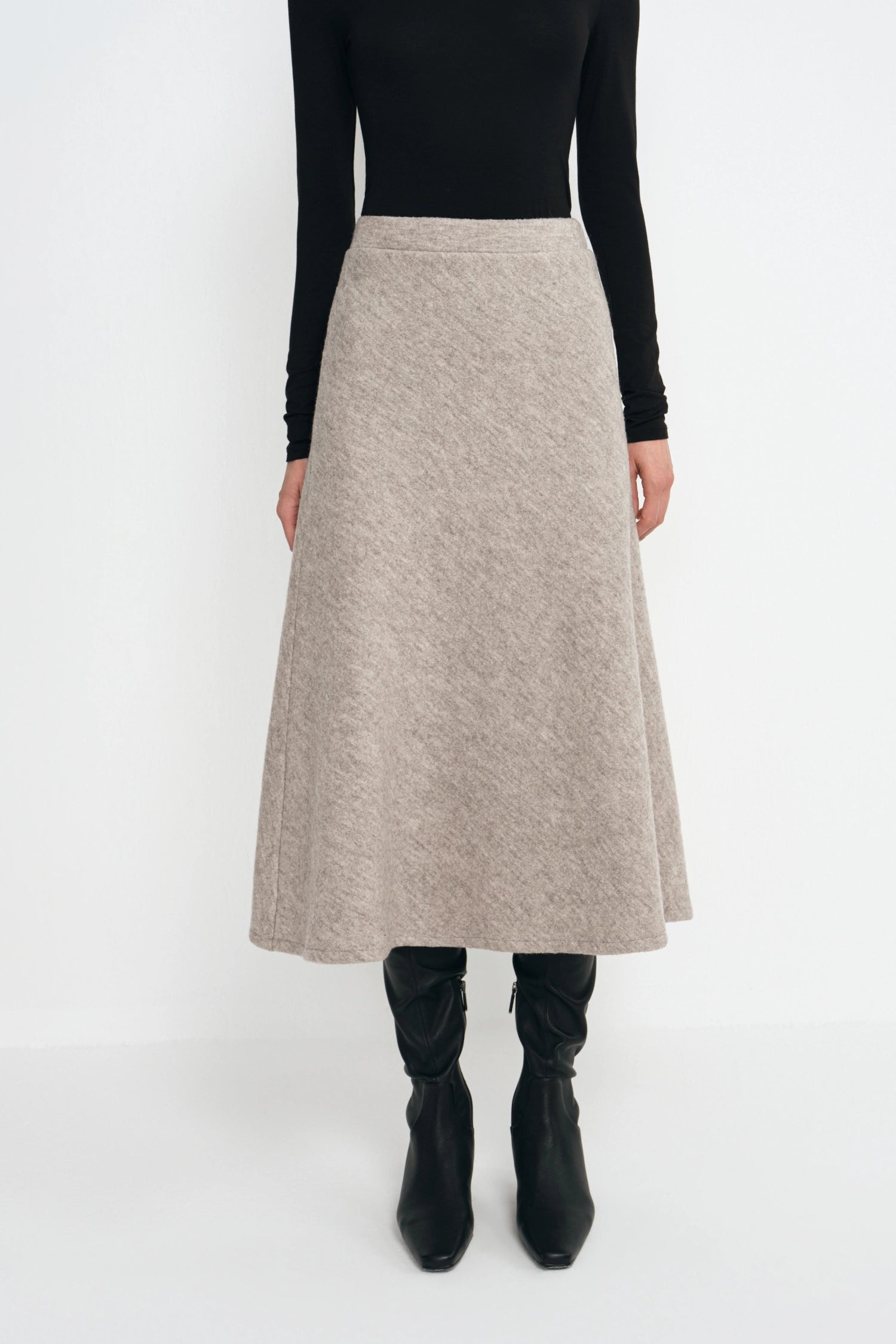 A model wearing a black long-sleeve top and a high-waisted, A-line midi skirt in a textured light beige or taupe knit. The model is also wearing black knee-high boots, photographed against a seamless white studio background.