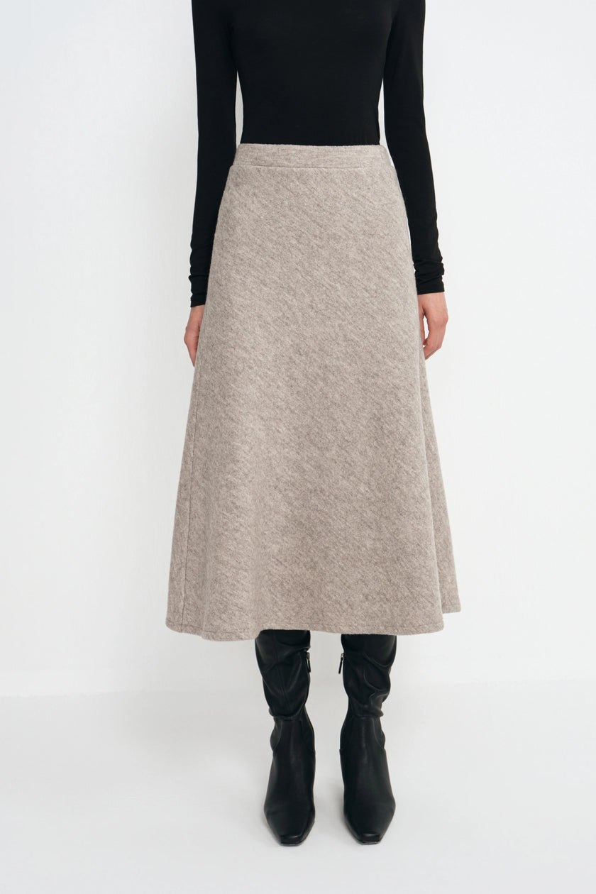 A model wearing a black long-sleeve top and a high-waisted, A-line midi skirt in a textured light beige or taupe knit. The model is also wearing black knee-high boots, photographed against a seamless white studio background.