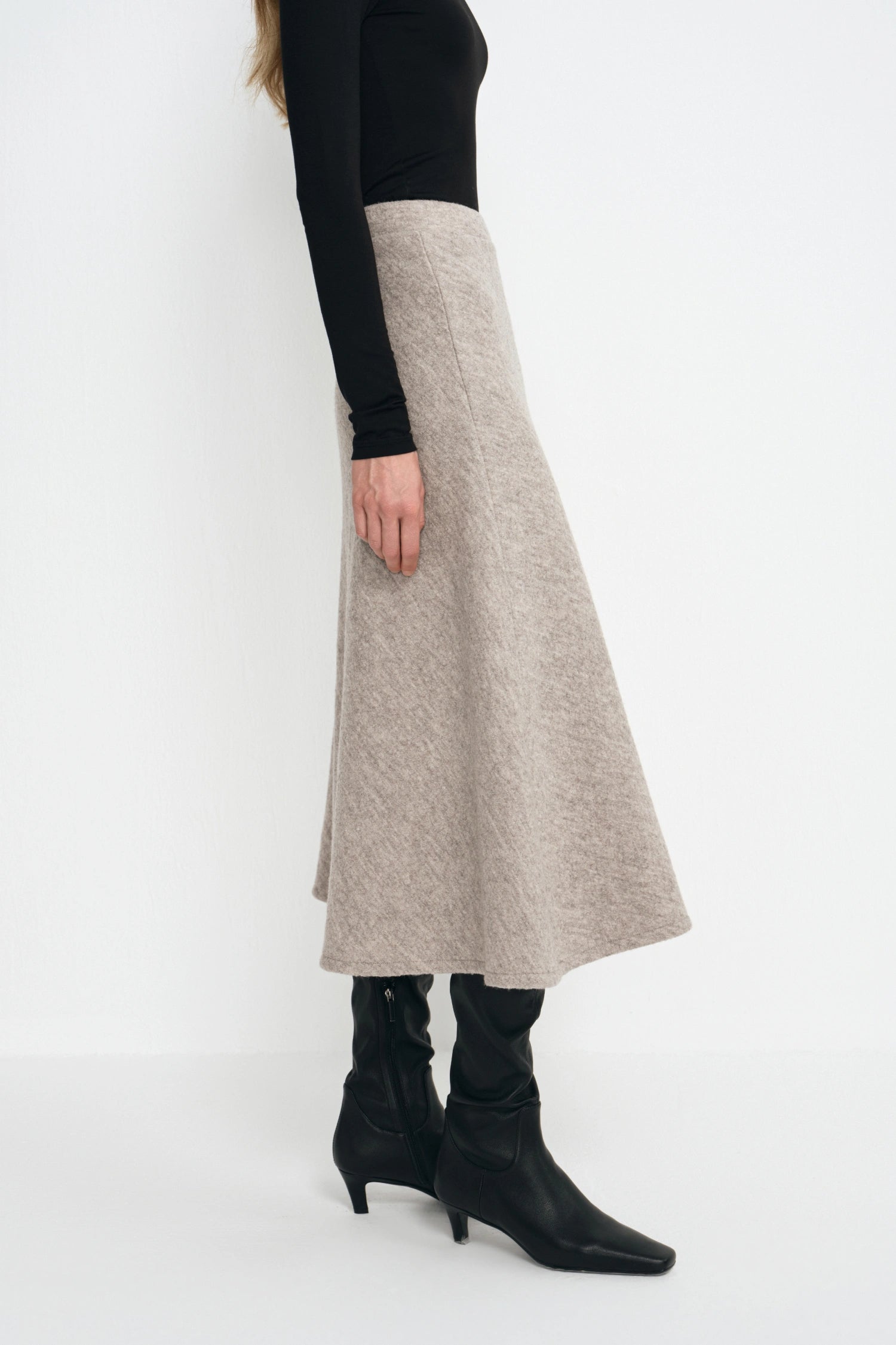 A model is shown from the side wearing a long, flared A-line midi skirt in a light oatmeal beige color, styled with a black long-sleeve top and black pointed-toe ankle boots.