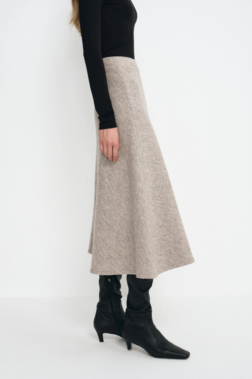 A model is shown from the side wearing a long, flared A-line midi skirt in a light oatmeal beige color, styled with a black long-sleeve top and black pointed-toe ankle boots.