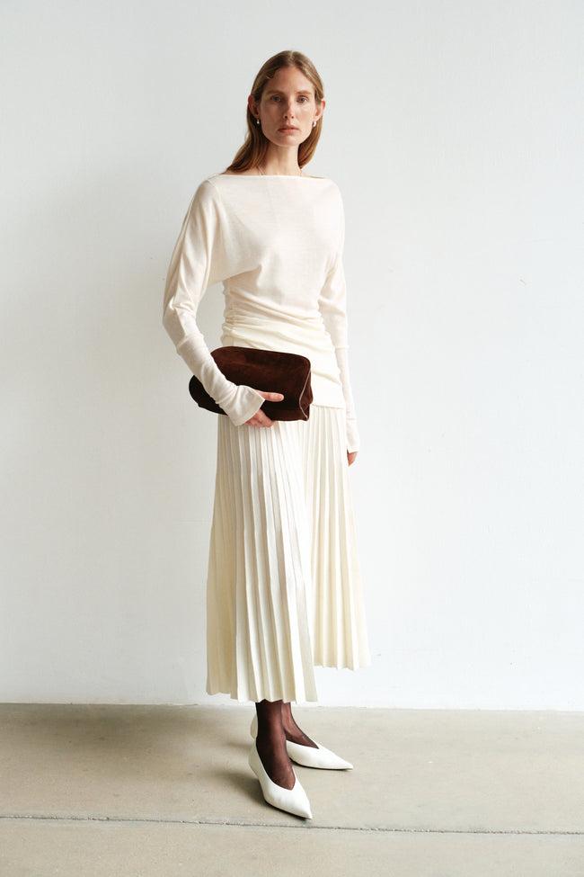 A model wearing the Amélie Accordion-Pleat Skirt in ecru, styled with a cream ruched long-sleeve top, brown clutch and white pointed shoes.