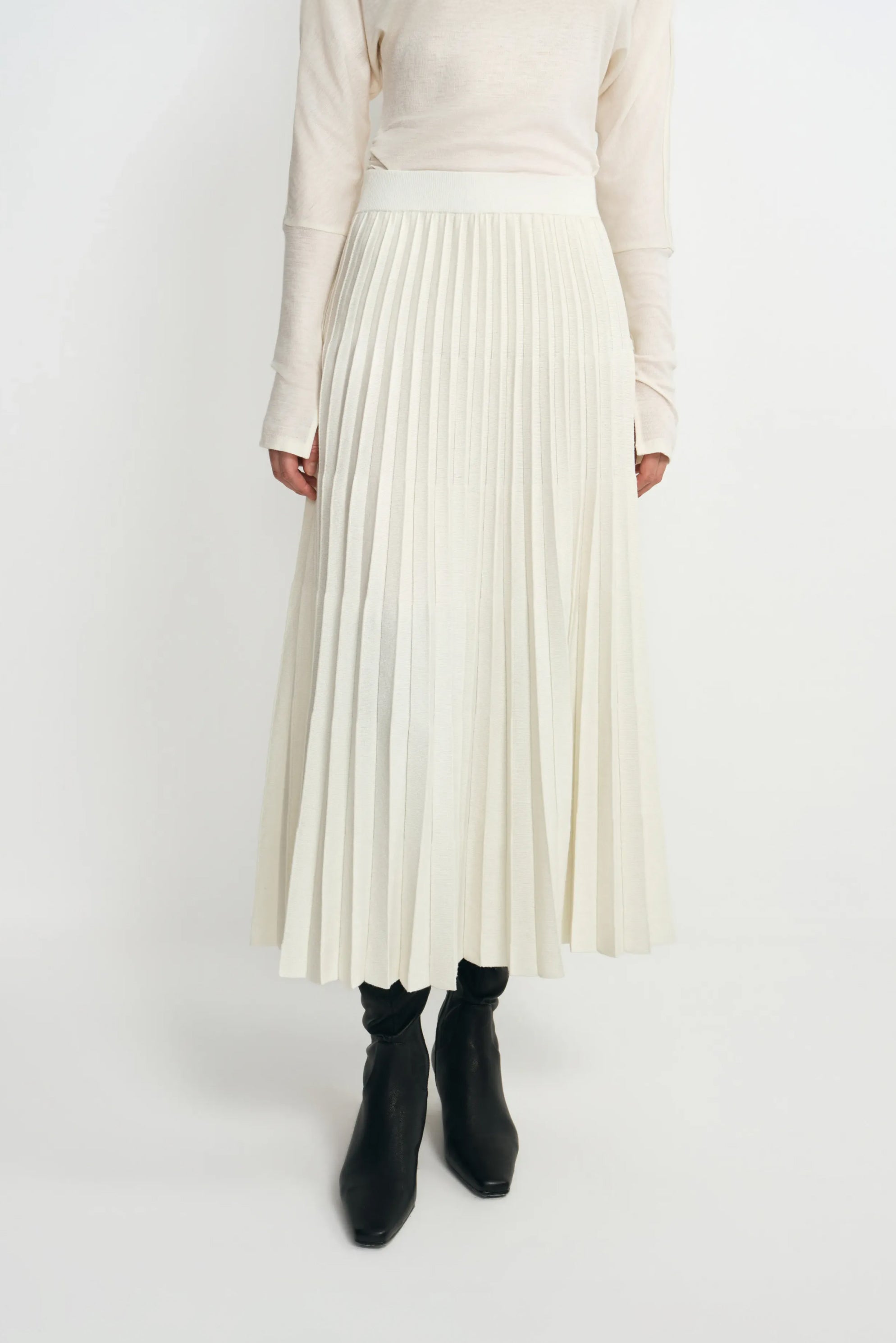 Front detail of an ecru wool-blend midi skirt on a model, showing the wide waistband and accordion pleats, paired with a tucked cream top and black boots.