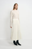 Three-quarter view of a model in an ecru accordion-pleated midi skirt, styled with a tucked cream boatneck top and black heeled boots.