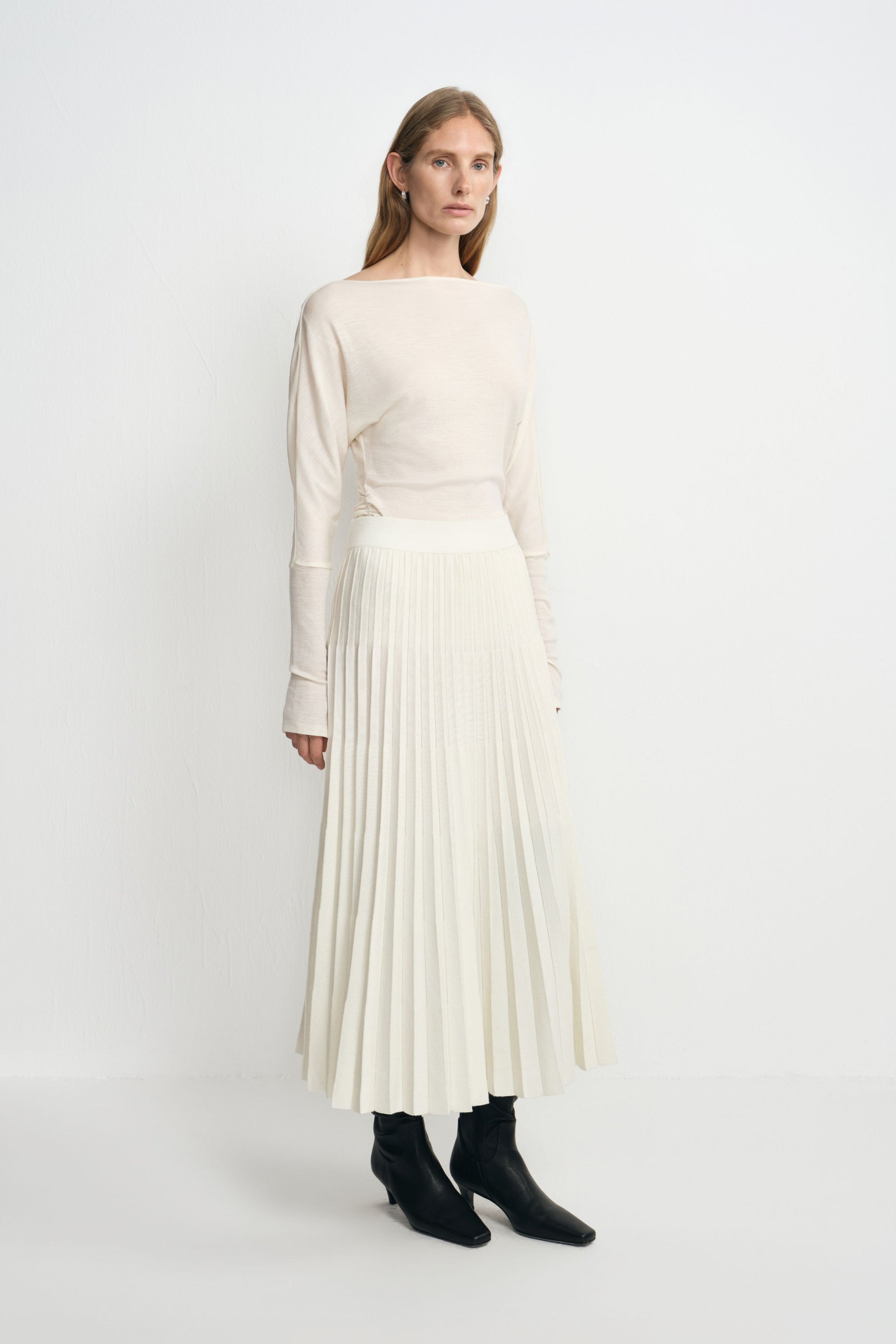 Three-quarter view of a model in an ecru accordion-pleated midi skirt, styled with a tucked cream boatneck top and black heeled boots.