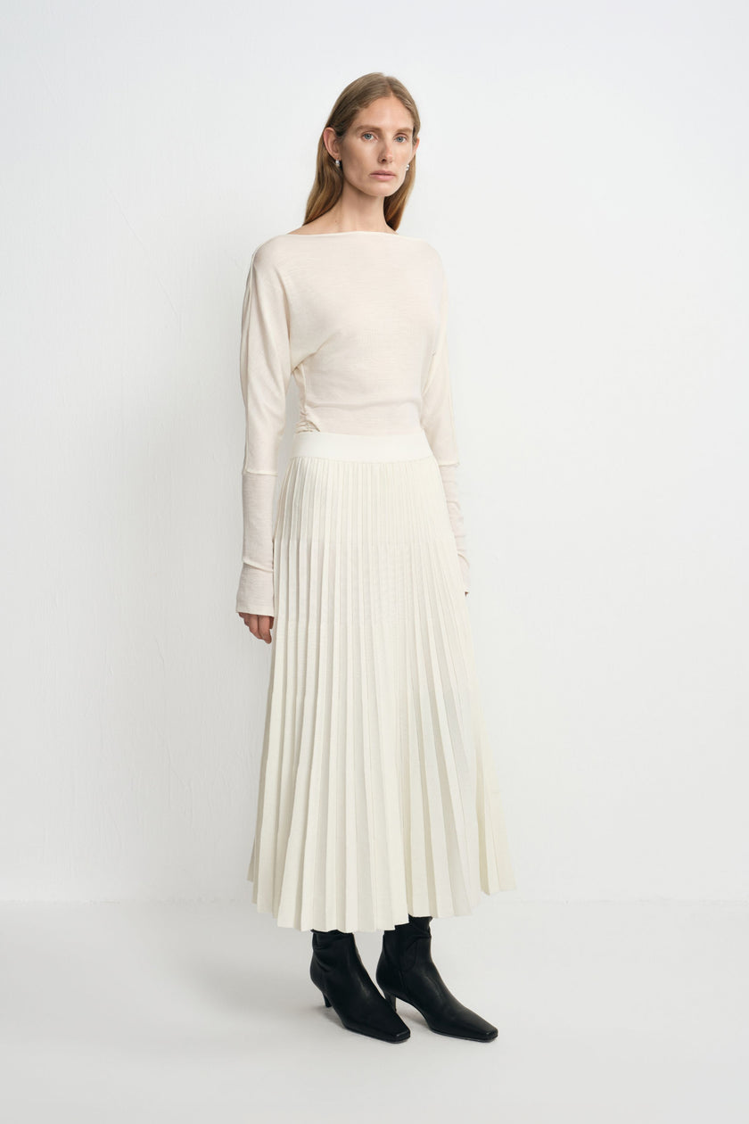 Three-quarter view of a model in an ecru accordion-pleated midi skirt, styled with a tucked cream boatneck top and black heeled boots.