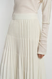 Close-up of an ecru wool-blend skirt on a model, showing the wide ribbed waistband and closely spaced accordion pleats beneath a tucked cream top.