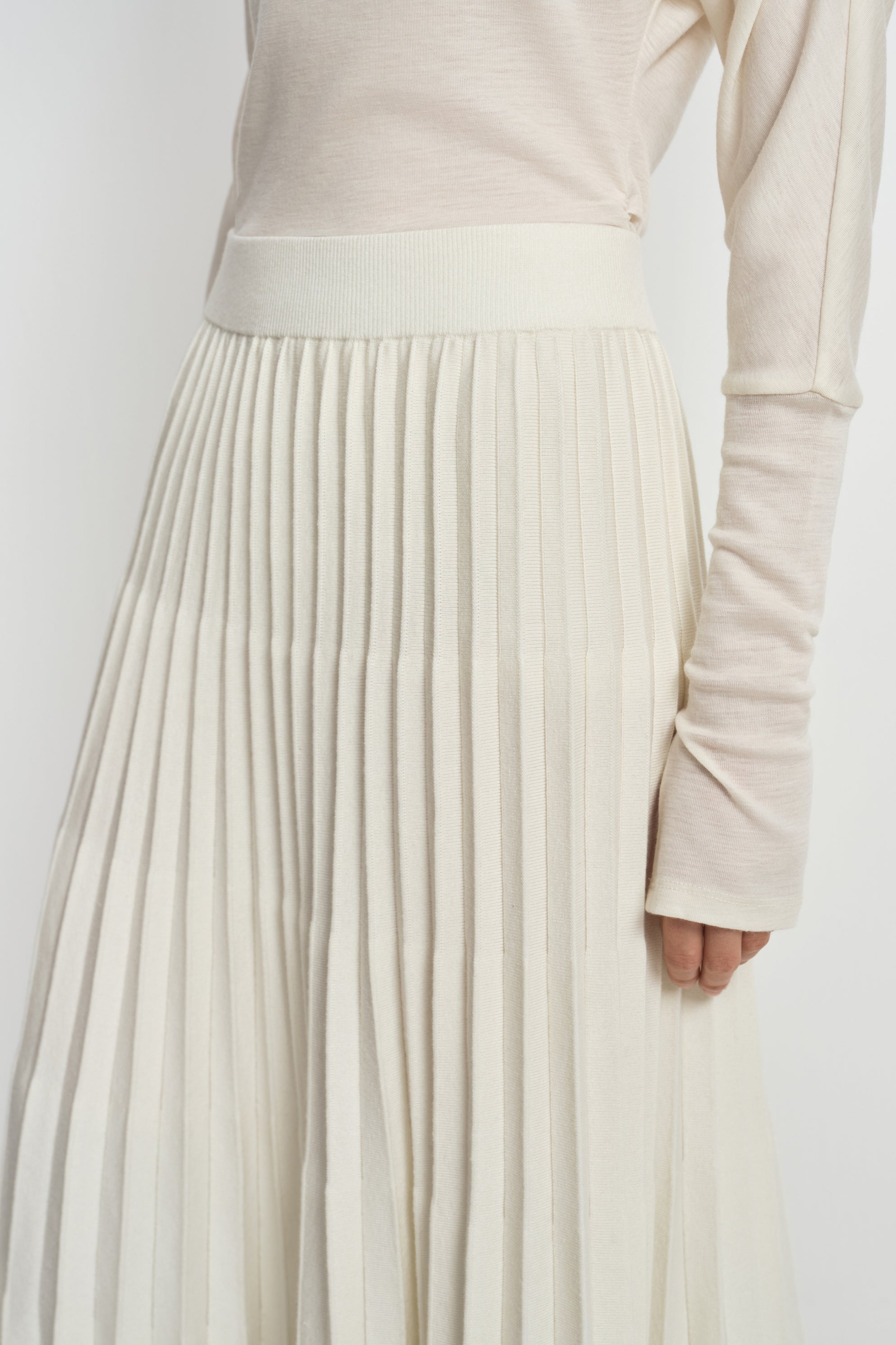 Close-up of an ecru wool-blend skirt on a model, showing the wide ribbed waistband and closely spaced accordion pleats beneath a tucked cream top.