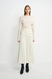 A model facing forward in an ecru pleated midi skirt, with narrow folds fanning toward the hem, paired with an untucked cream top and black boots.