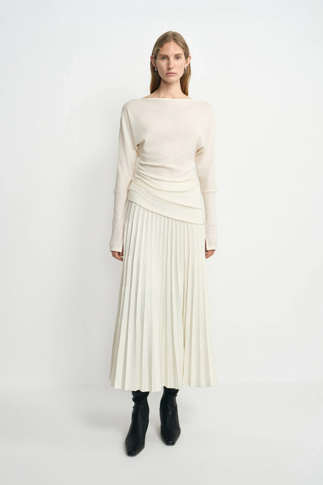 A model facing forward in an ecru pleated midi skirt, with narrow folds fanning toward the hem, paired with an untucked cream top and black boots.
