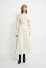 A model facing forward in an ecru pleated midi skirt, with narrow folds fanning toward the hem, paired with an untucked cream top and black boots.
